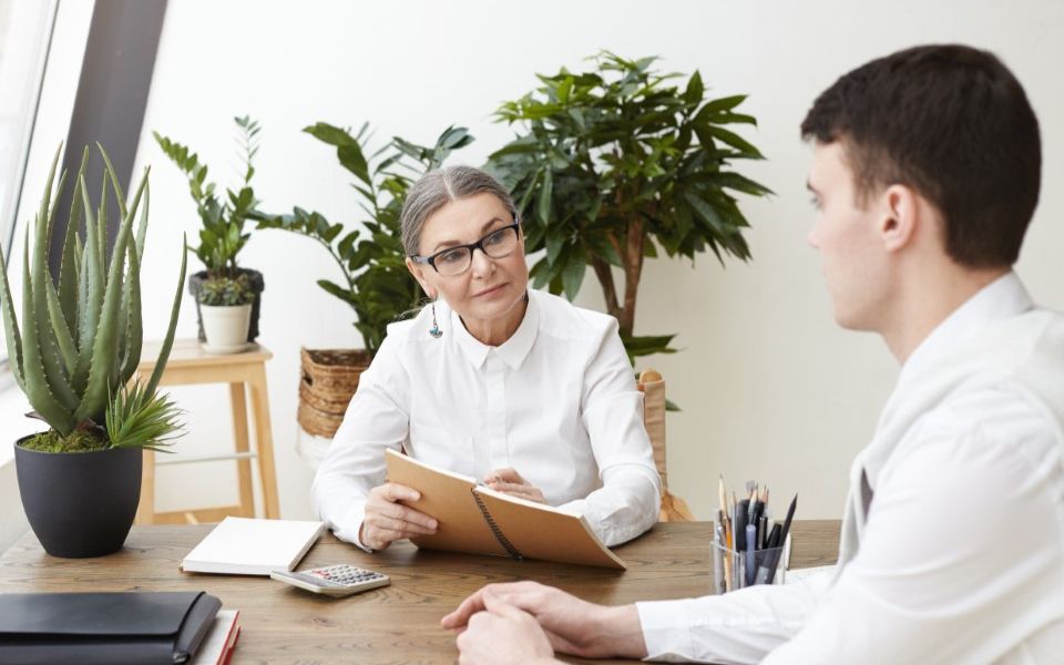 Dos personas sentadas a una mesa conversan. La mujer sostiene diferentes papeles con actitud de buscar respuestas, como en una entrevista laboral o de orientación al empleo.