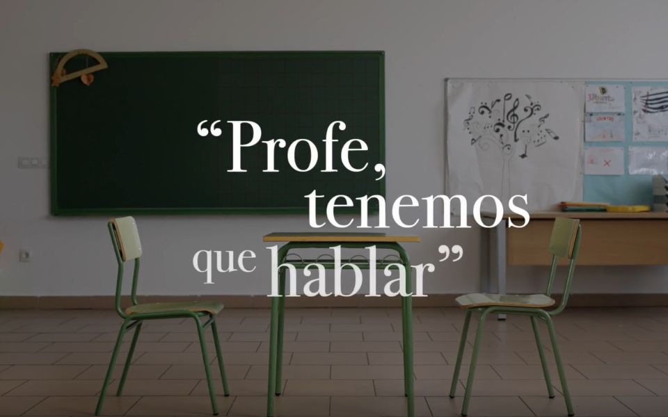 Fotografía de una clase con un pupitre verde y dos sillas a los lados en primer plano, una pizarra verde al fondo. Sobreimpresionado en la imagen el texto: Profe, tenemos que hablar. Iniciativa contra el acoso escolar en el alumnado con autismo.