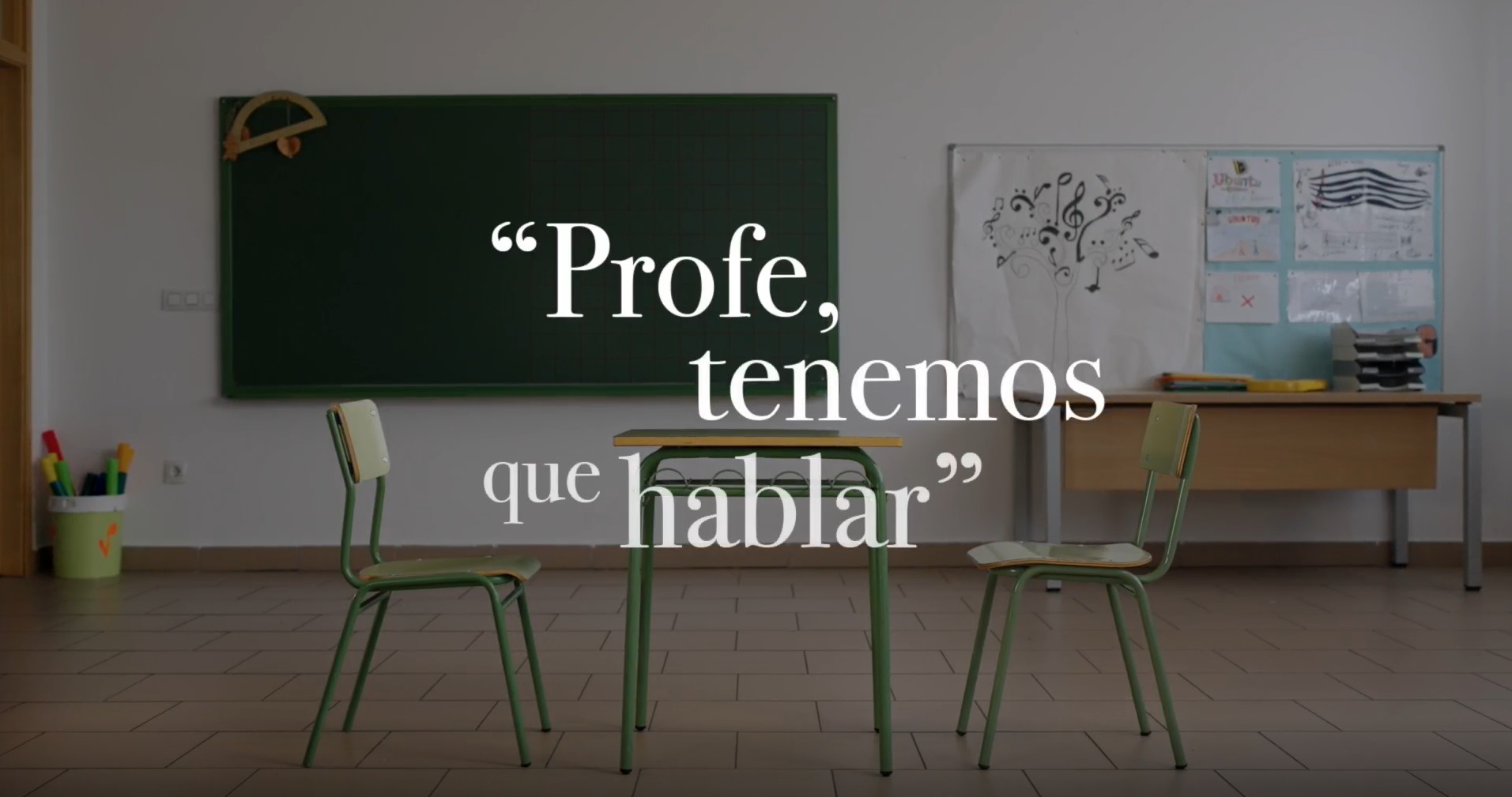 Fotografía de una clase con un pupitre verde y dos sillas a los lados en primer plano, una pizarra verde al fondo. Sobreimpresionado en la imagen el texto: Profe, tenemos que hablar.
