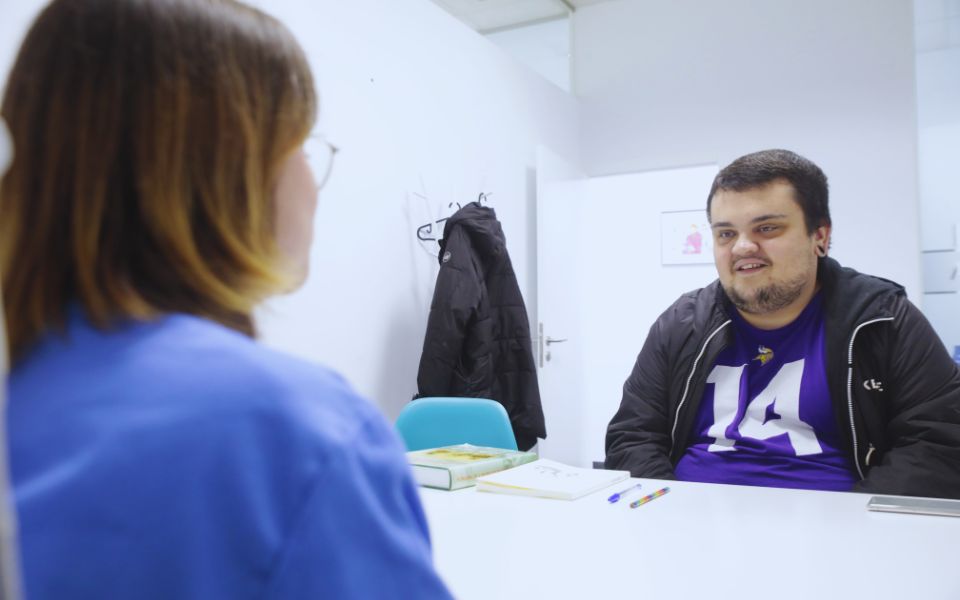 Fotografía de una persona tras una mesa hablando con una profesional del ámbito de la discapacidad.