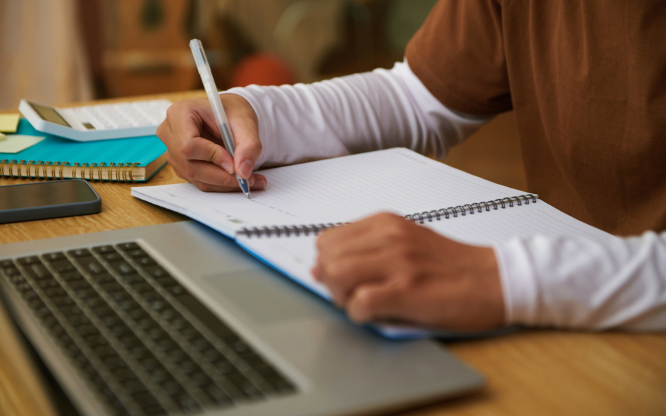 Una persona escribe en una libreta mientras mira un ordenador portátil